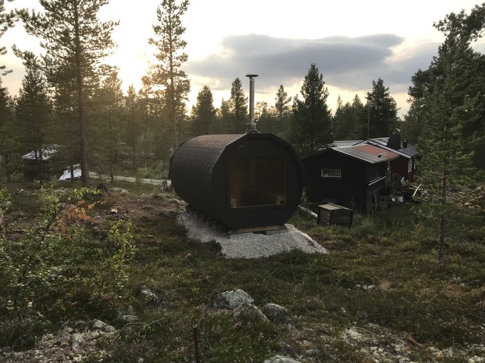 En svartmålad bastutunna med stora fönster och skorsten placerad i fjällterräng nära ett högt träd.