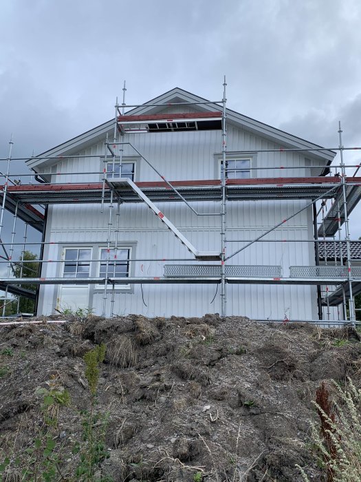 Ett hus under renovering med ställningar runt fasaden och nymålad övervåning mot en molnig himmel.