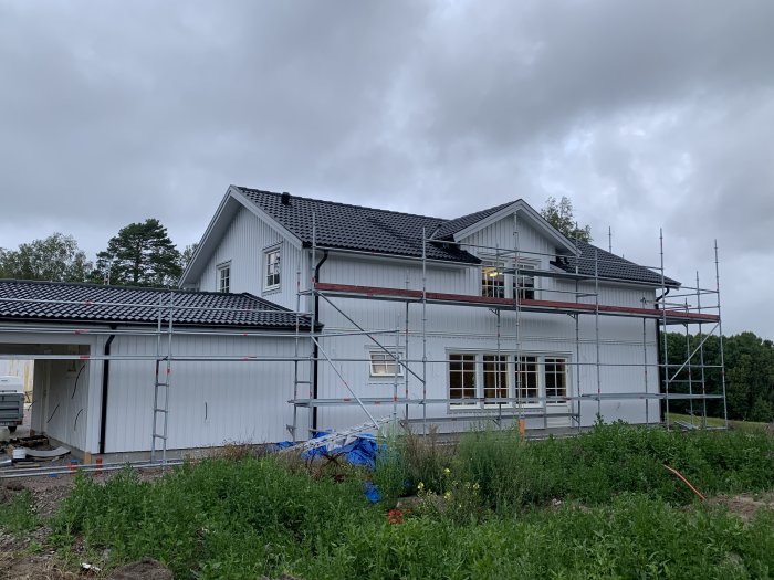 Vitmålat hus under renovering med ställningar runtom, taket färdigt och färgarbete pågår på bottenvåningen.