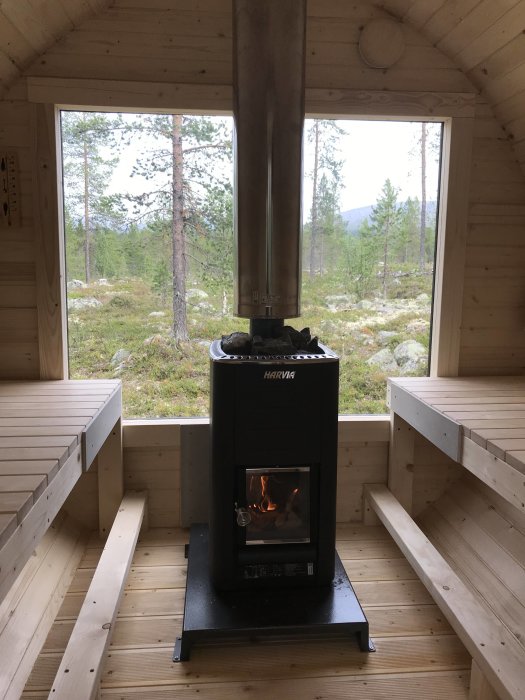 Interiör av en bastu med stor fönstervy över fjällandskap, vedeldad bastuugn tänd, träbänkar.