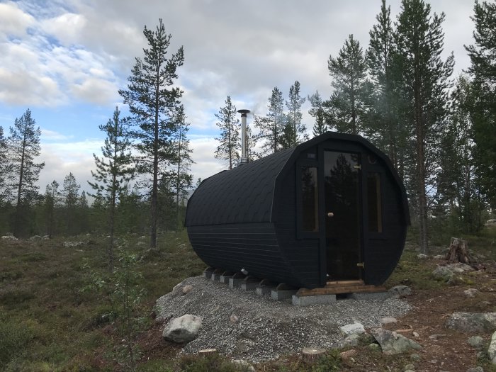 Svartmålad bastu med större fönster i en skogsmiljö, monterad på grusunderlag i Fjällvärlden.