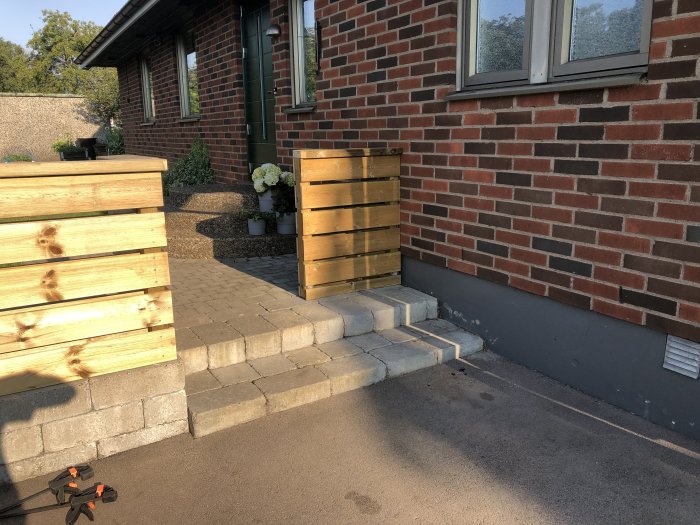 Träplank och en öppen grind i trä framför en tegelhusfasad och stensatt uppfart.