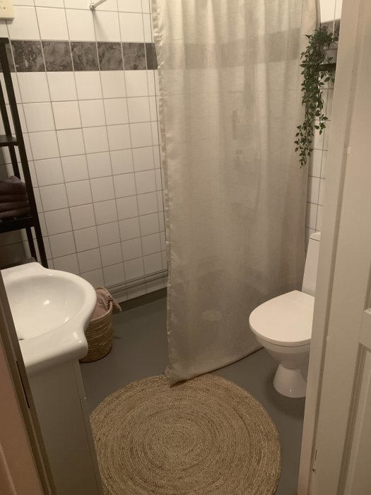 Renoverat badrum med målad golvbeläggning, vita kakelväggar, duschdraperi och badrumsmattor.