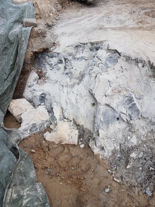 Stor stenblock i en grävning, delvis täckt av en presenning, med mindre stenar och grus.