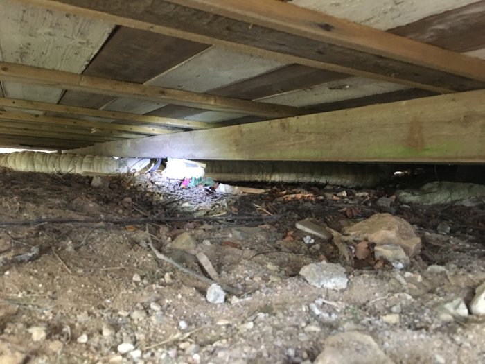 Vy under ett hus med bjälkar, isolering och ojämnt placerade stödplintar och stenar på marken.