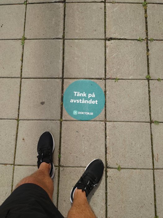 Person står på trottoar med skylt som uppmanar att "Tänka på avståndet", sponsrad av doktor.se.