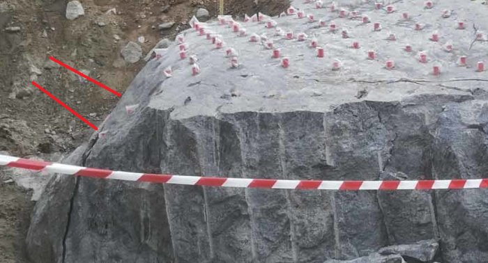 Granitberg med borrade hål förberedda för sprängning, markerade med rödvita varningssnören och dynamithattar.