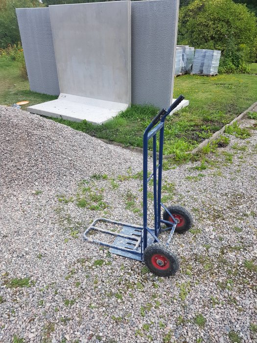 Blå säckkärra på grus framför betongplatta och gråa betongväggar i trädgårdsmiljö.
