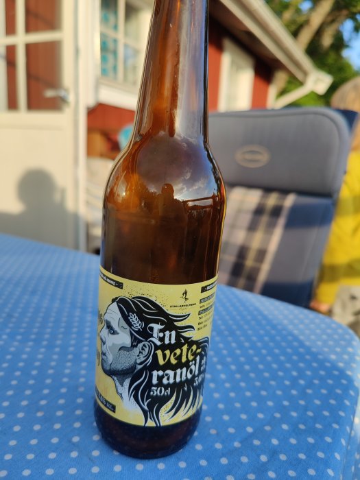 Flaska med veteöl på ett blåprickigt bord utomhus, etiketten visar en illustrerad mansprofil.
