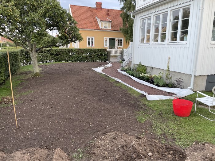 Nygrävd gång med utlagd markduk i trädgård framför hus, med träd, grässvål och byggmaterial synligt.