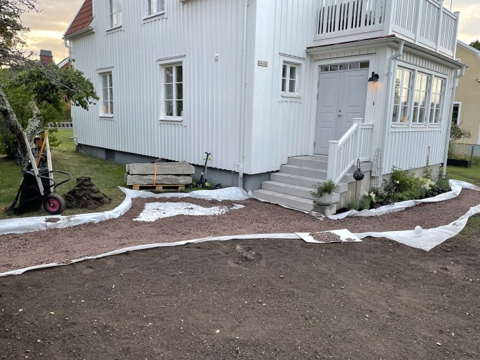 Förberedning av gångväg i trädgård med markduk, bärlager av stenmaterial och utspridd matjord, nära vit villa.