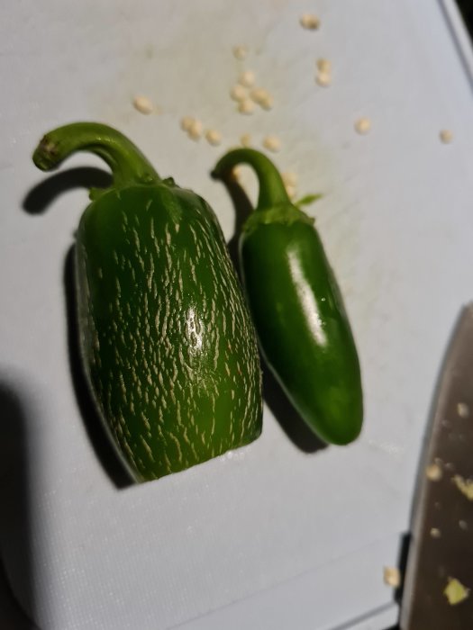 Två gröna jalapeño-chilifrukter med ränder på ett skärbräde bredvid några frön och en kniv.