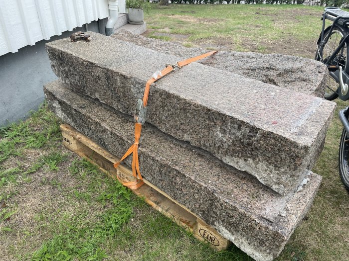 Fyra gråa granitblocksteg staplade på en pall, bundna med orange spännband, bredvid en cykel utomhus.