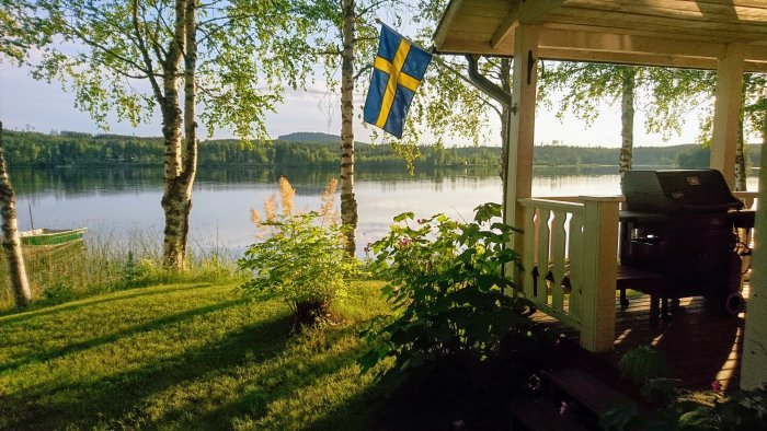 Veranda med svensk flagga framför en sjöutsikt, grill och grönska i sommarljus.