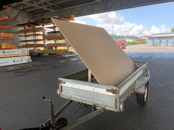 En stor MDF-skiva lastad på en släpvagn under ett tak med byggnadsmaterial i bakgrunden.