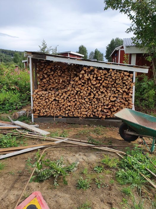 Vedhög av klyvt lärkträ under tak, med röda lador och grönska i bakgrunden, och en grön skottkärra i förgrunden.