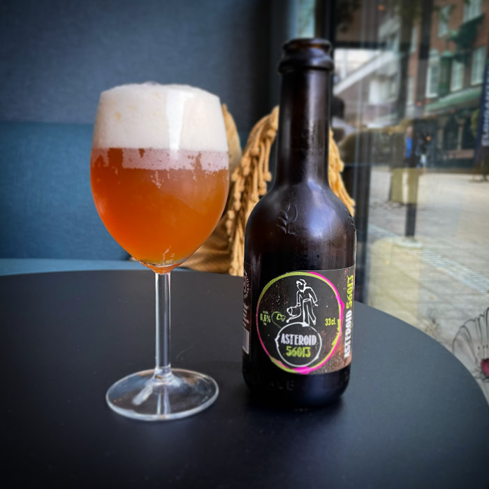 Ett glas fyllt med öl bredvid en ölflaska med etiketten "ASTERBROID 56013" på ett svart bord.