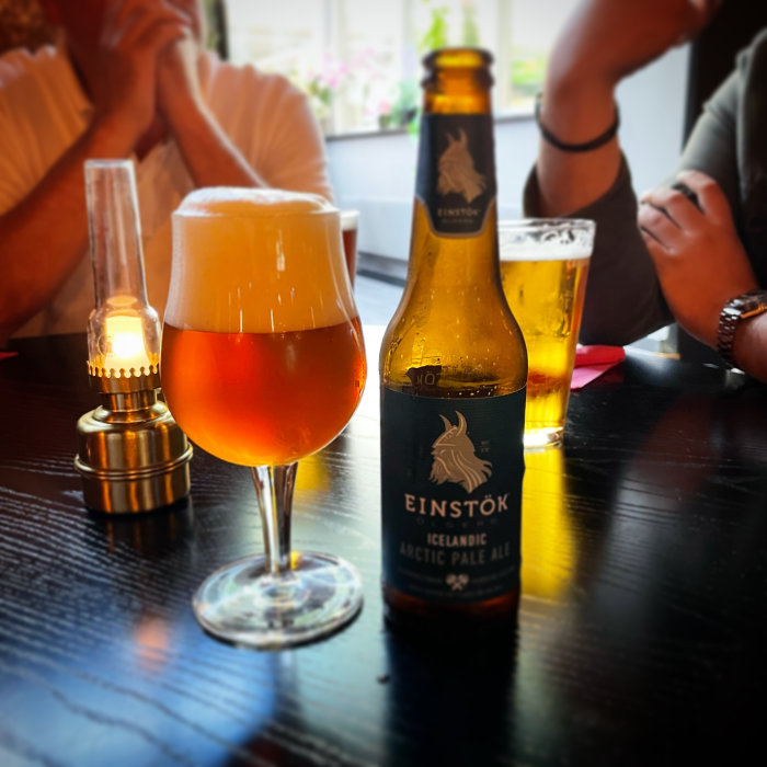 Flaska och glas av Einstök öl på bord med lampa och okänsliga personer i bakgrunden.