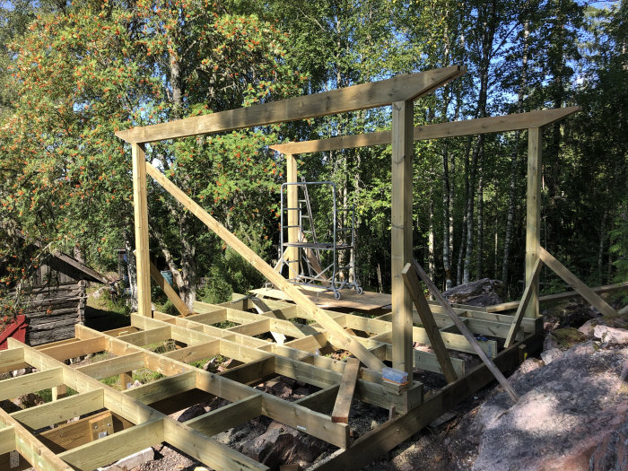 Pågående byggnation av en träaltan med pergola, visar stomme och regelverk i ett skogsnära område.