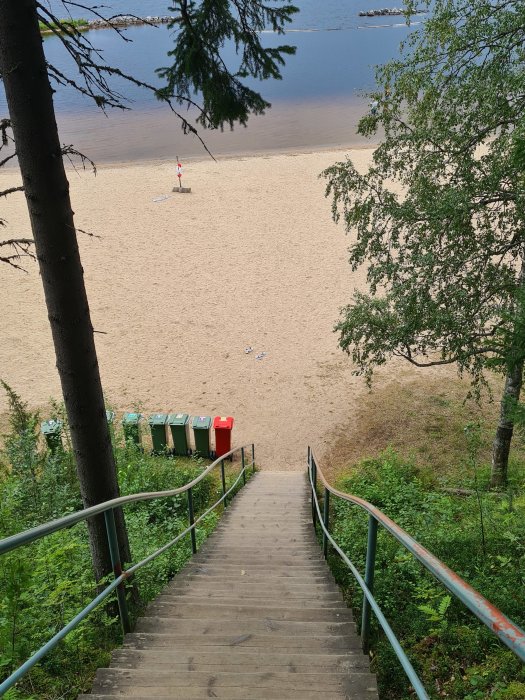 Trätrappa som leder ner till en sandstrand med soptunnor vid sidan och en sjö i bakgrunden.