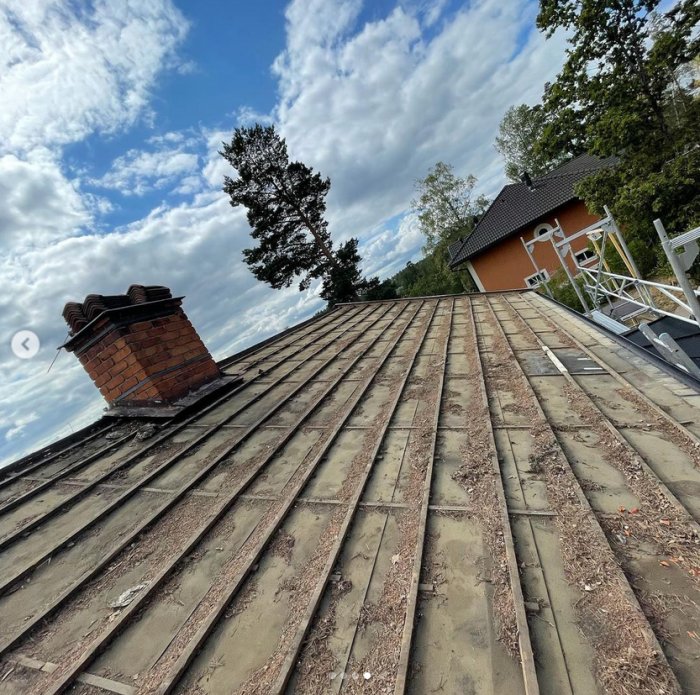 Renovering av gammalt tak med tegelpannor och råspont, byggsäckar syns, samt utsikt mot skorsten och hus.