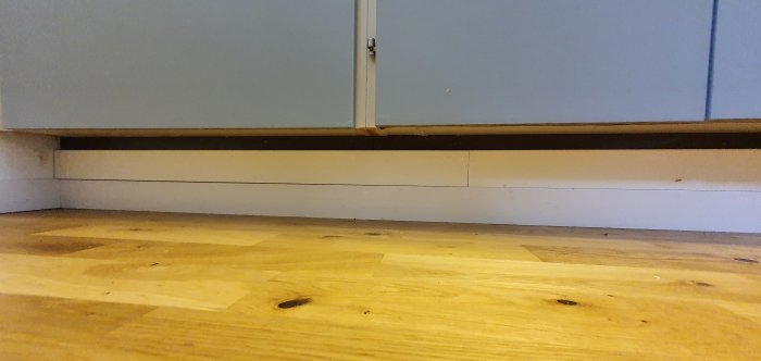 Fotografi som visar köksskåpens botten med en fast spikad sockellist, mot en träparkettgolv.