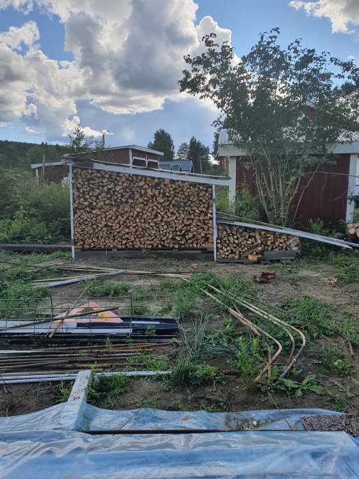 Stapel av klyvd lärkved i vedupplag mot en himmel med moln, med byggmaterial i förgrunden.