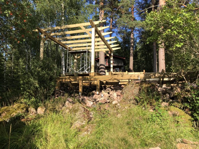 Nytt trädäck under konstruktion med tjocka bjälkar och reglar i en skogsmiljö vid en stuga.