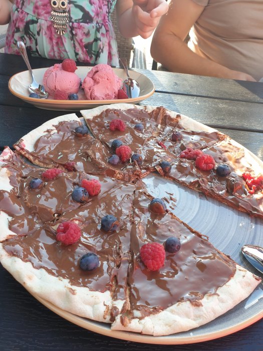 Dessertpizza med smält choklad och bär samt en tallrik med rosa glass på ett bord.