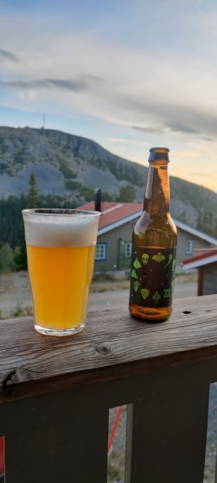 Ölflaska och ölglas på balkongräcket med en bergsutsikt i bakgrunden vid skymningen.
