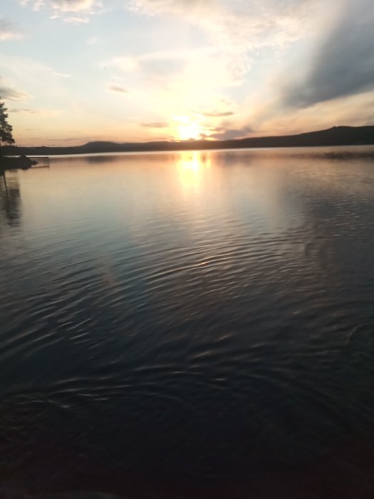 Solnedgång över en lugn sjö med reflekterande solstrålar och siluett av berg i bakgrunden.