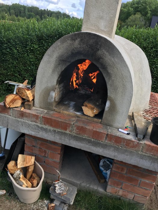 Hemgjord tegel vedeldad pizzaugn med brinnande ved och verktyg på sidan.