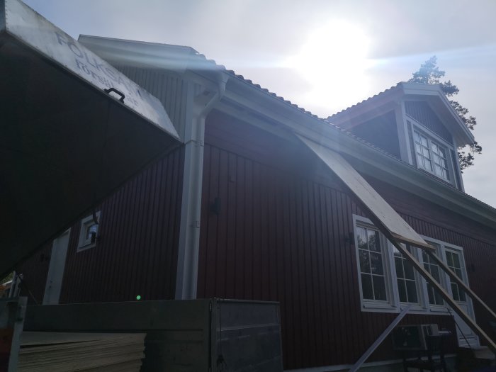 En hemgjord skivline för uppföring av plywoodskivor till taket på ett rödmålat hus med vit fönsterkarm.