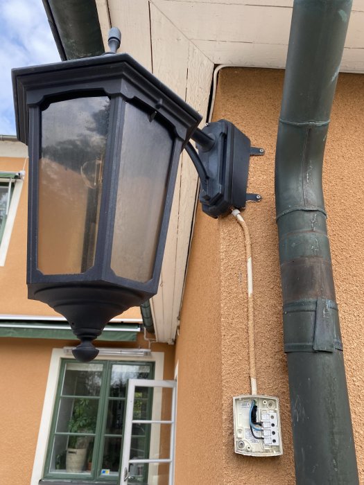Öppen utomhusarmatur vid husvägg med synlig anslutning och rör som går ned till en sensor vid stående stuprör.