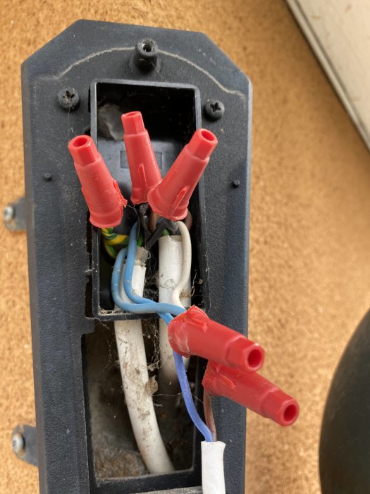 Öppen armatur med elektriska anslutningar och röda kopplingsklämmor, mot en bakgrund av rör och spindelväv.