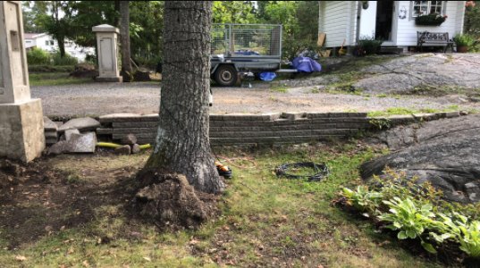 Delvis rivet stenmurprojekt vid en högre belägen bilparkering intill gräsmatta med verktyg och material synliga.