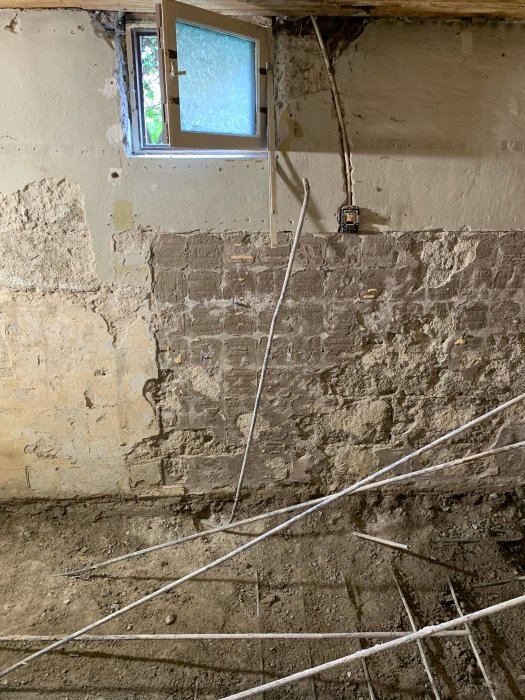 Nedbilat golv i källarbadrum med synliga armeringsjärn, rör och murat väggparti under renovering.