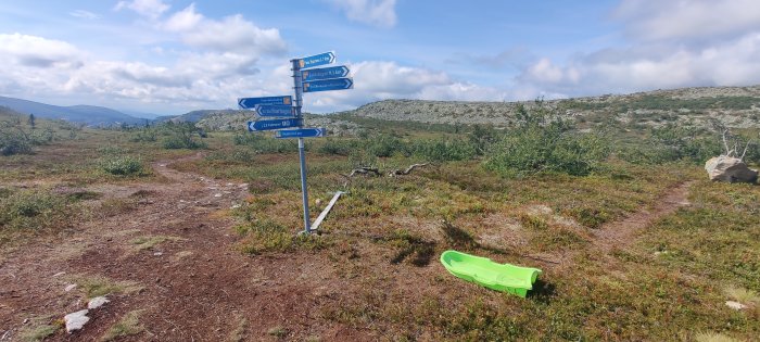 Vägvisarskyltar i fjällterräng med en grön pulka liggande i marken.