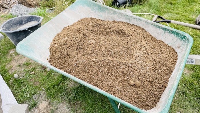 En skottkärra fylld med gjutgrus på en gräsmatta, nära en svart hink och en spade.