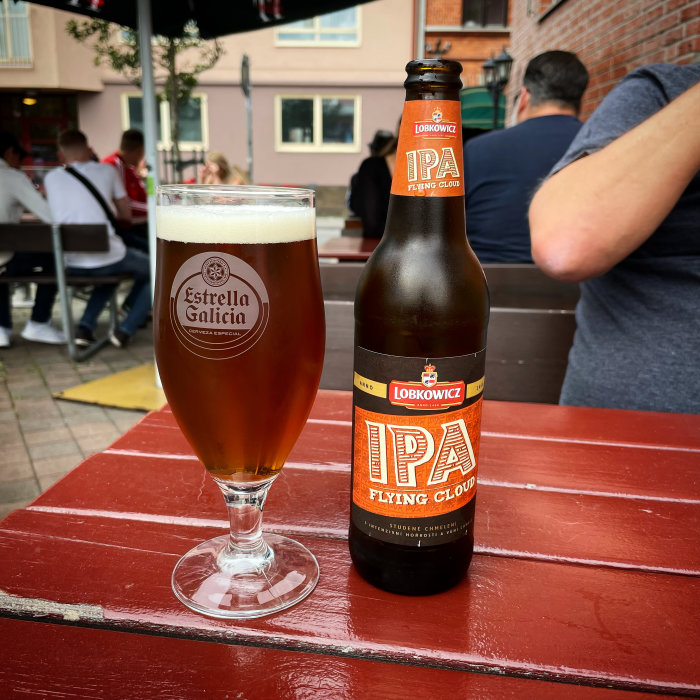 En IPA-flaska bredvid ett glas öl på ett rött bord utomhus med restauranggäster i bakgrunden.