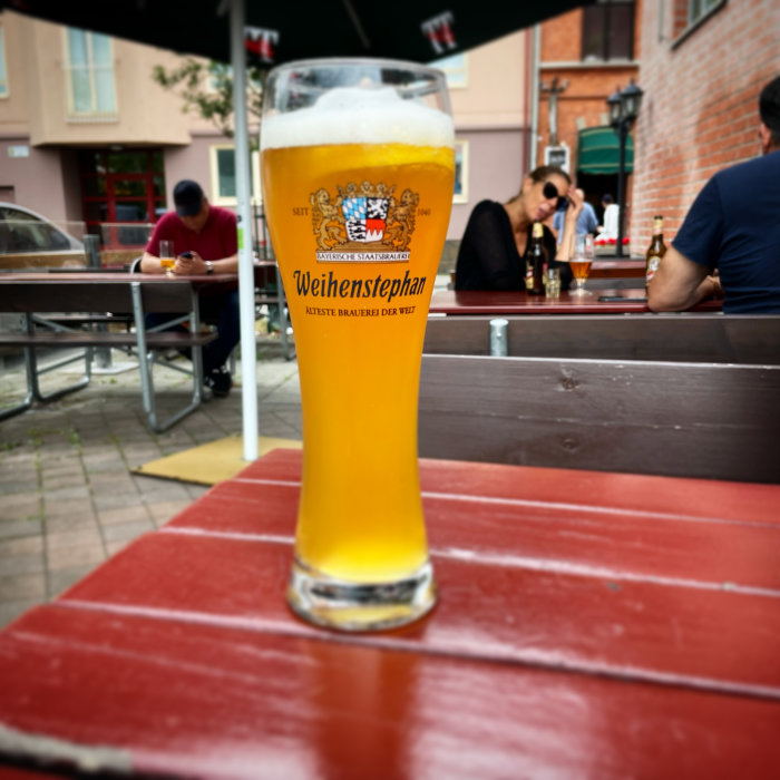 Ett glas Weihenstephaner öl på ett utomhusbord med suddiga personer i bakgrunden.