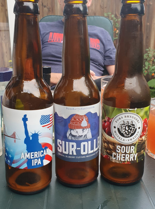 Tre ölflaskor med etiketter "AMERICAN IPA", "SUR-OLLE" och "SOUR CHERRY" framför en suddig person.