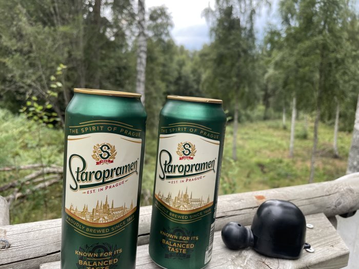 Två burkar Staropramen öl på en träbänk utomhus med skog i bakgrunden.