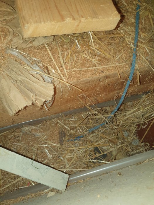 Loft med sågspån och hörester på träbjälklag samt rör och plastduk i ekonomibyggnad.