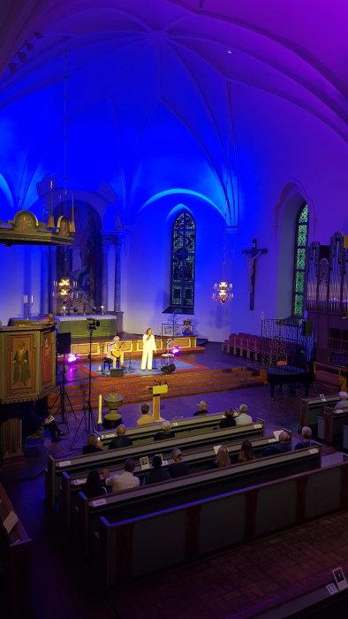 Interiör av kyrka med blå belysning, personer i bänkar och en person som sjunger med gitarrist.