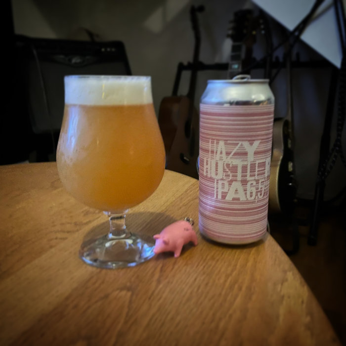 Ett glas med grumlig öl bredvid en burk IPA och en liten rosa grisfigur på ett träbord.