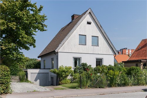 Ett äldre hus från 1924 med vit panel på fasaden och ospröjsade fönster omgivet av grönska.