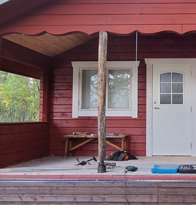 Renoverad domkraft och gängstift under trästolpe på ett rödmålat hus med veranda.