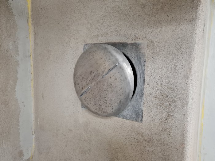 Använt utseende av en rund tallriksventil i aluminium infälld i en murstock, med synliga missfärgningar och skador.