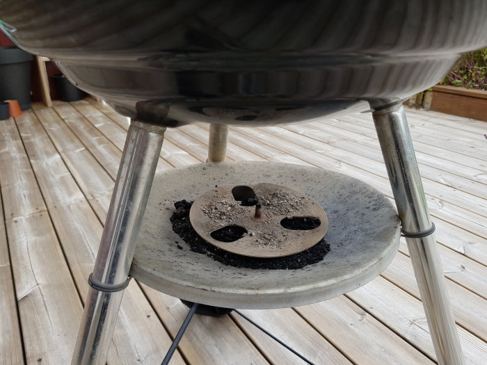 Webergrill med lossnad bottenventil och aska som ligger på en stenplatta på en trädäck.